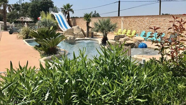 A pool area with a water slide, lounge chairs, and a stone wall. - Camping 4 étoiles - Piscine  - ccbgce0 (Sorgues)