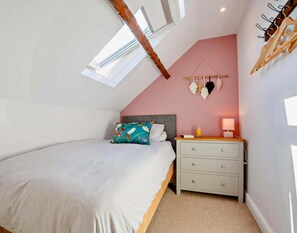 A bedroom with a sloped ceiling, a bed with white bedding, a nightstand with a lamp, and a wall-mounted macrame decoration. - Cottage Sleeps 5 With Parking and Garden (Amlwch)