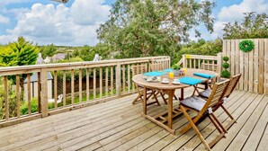 Eine Holzterrasse mit einem runden Tisch und Stühlen, ein Holzzaun und Blick auf Bäume und Grün.