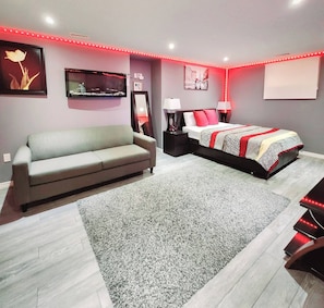 A modern bedroom with a grey sofa, a large bed with patterned bedding, a grey carpet, and red LED strip lighting. - Staycation Studio Apartment (London)