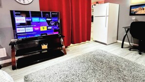 A living room with a flat-screen TV, a clock, a red curtain, a refrigerator, and a carpet. - Staycation Studio Apartment (London)