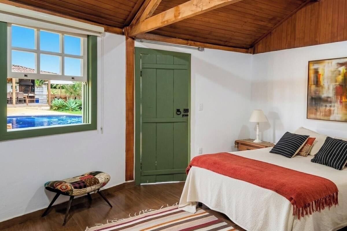 Une chambre à coucher comprenant un lit, une table de chevet, une lampe, un tableau, une fenêtre donnant sur une piscine et une porte.