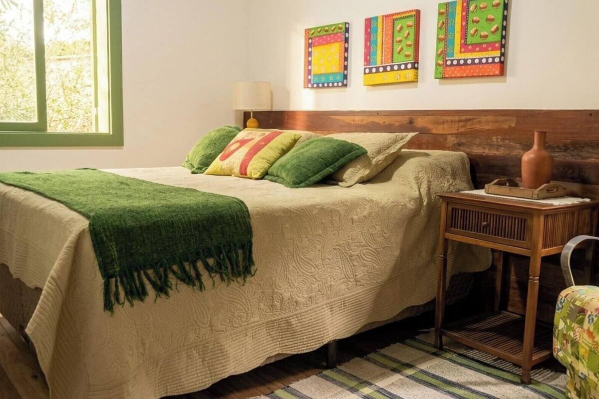 Un lit recouvert d’une couverture verte, une table de chevet en bois avec un vase, et une fenêtre donnant sur des arbres.