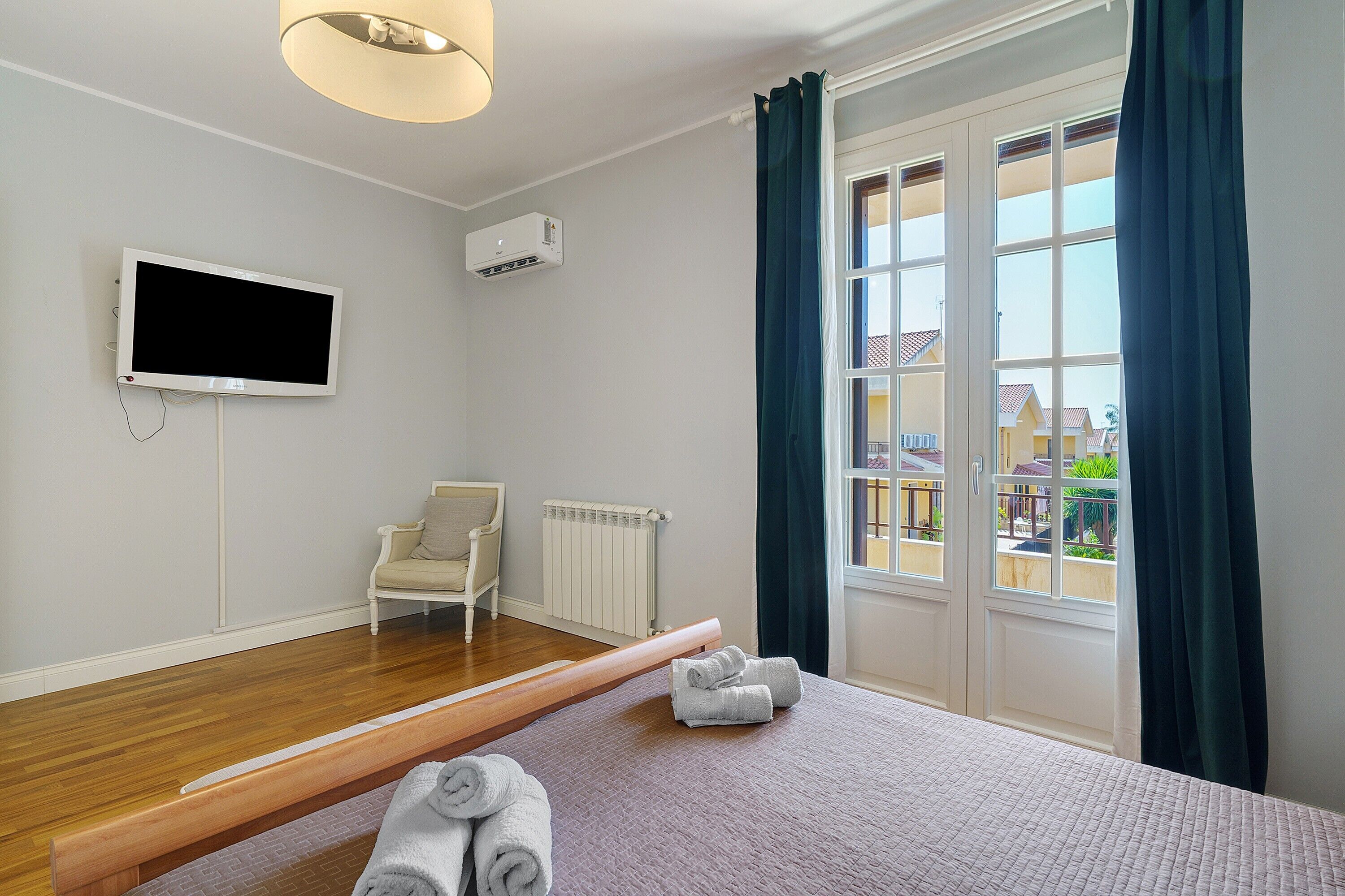 Ein Hotelzimmer mit Bett, Sessel, Fernseher, Klimaanlage, einem Fenster mit Vorhängen und Blick auf umliegende Gebäude.