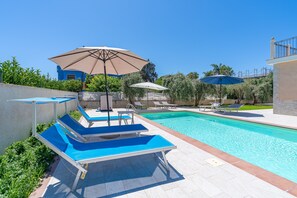 Una piscina con sillas de descanso y sombrillas, rodeada de vegetación y un edificio.