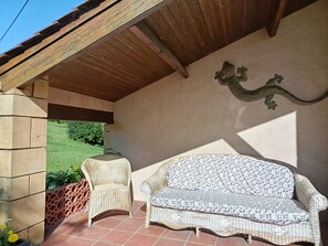 A covered patio area with a wicker sofa and chair, a decorative wall piece, and a view of greenery outside. - "Gîte des Cerisiers" Apartment with Pool, Terrace and Private Garden (Saint-Germain-de-Belvès)