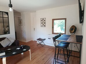 A room with a wooden floor, a table with chairs, a sofa, a desk with a chair, a window, and a wall-mounted fan. - "Gîte des Cerisiers" Apartment with Pool, Terrace and Private Garden (Saint-Germain-de-Belvès)