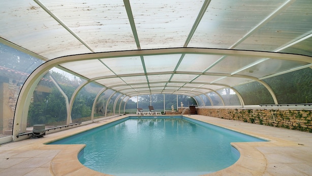 A covered swimming pool with a curved edge, a stone wall, and a view of greenery outside.
