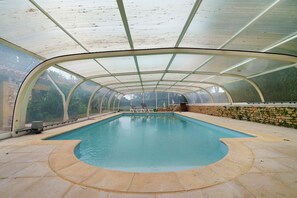 Ein überdachter Pool mit geschwungener Kante, einer Steinwand und Blick ins Grüne.