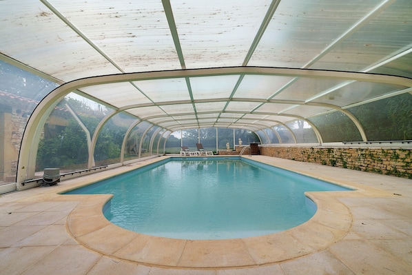 A covered swimming pool with a curved edge, a stone wall, and a view of greenery outside.