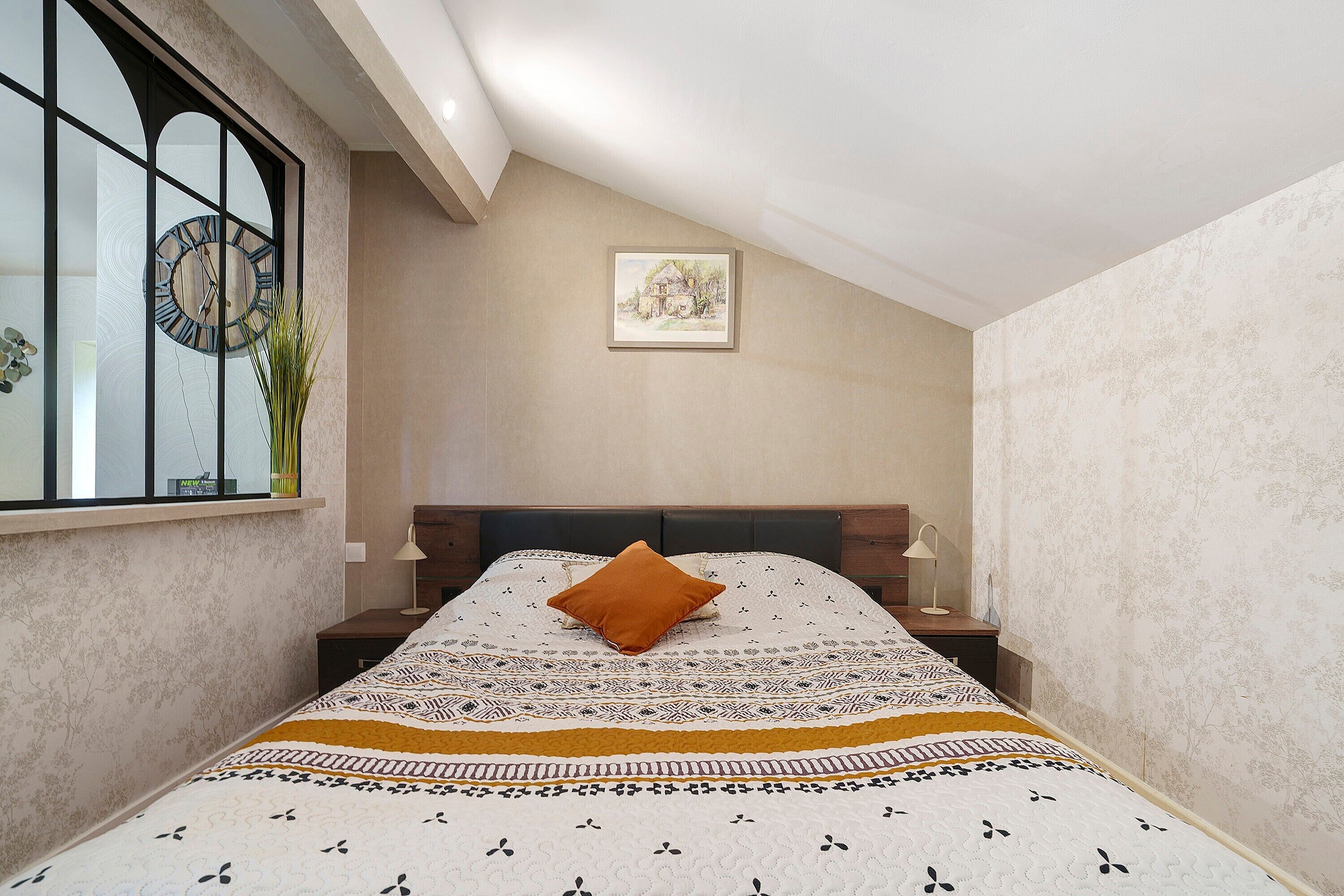 A bedroom with a bed, a nightstand, a lamp, a window with a clock, and a wall-mounted picture.