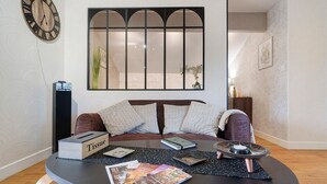 A living room with a sofa, a round table with magazines, a clock, and a window with a view.