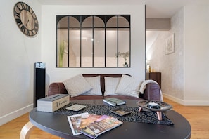 A living room with a sofa, a round table with magazines, a clock, and a window with a view.
