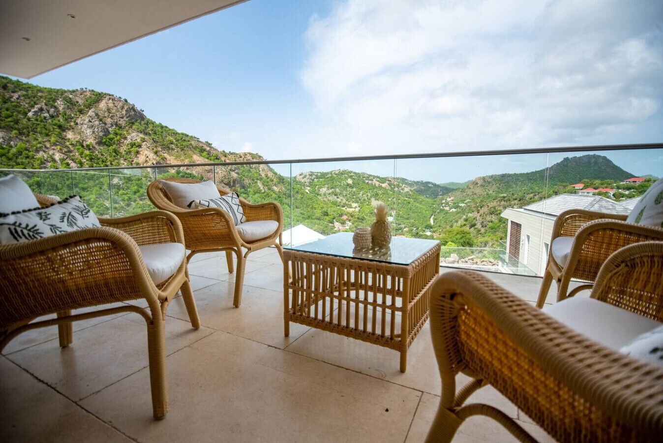 A balcony with wicker furniture, a glass table, and a scenic view of a mountainous landscape.