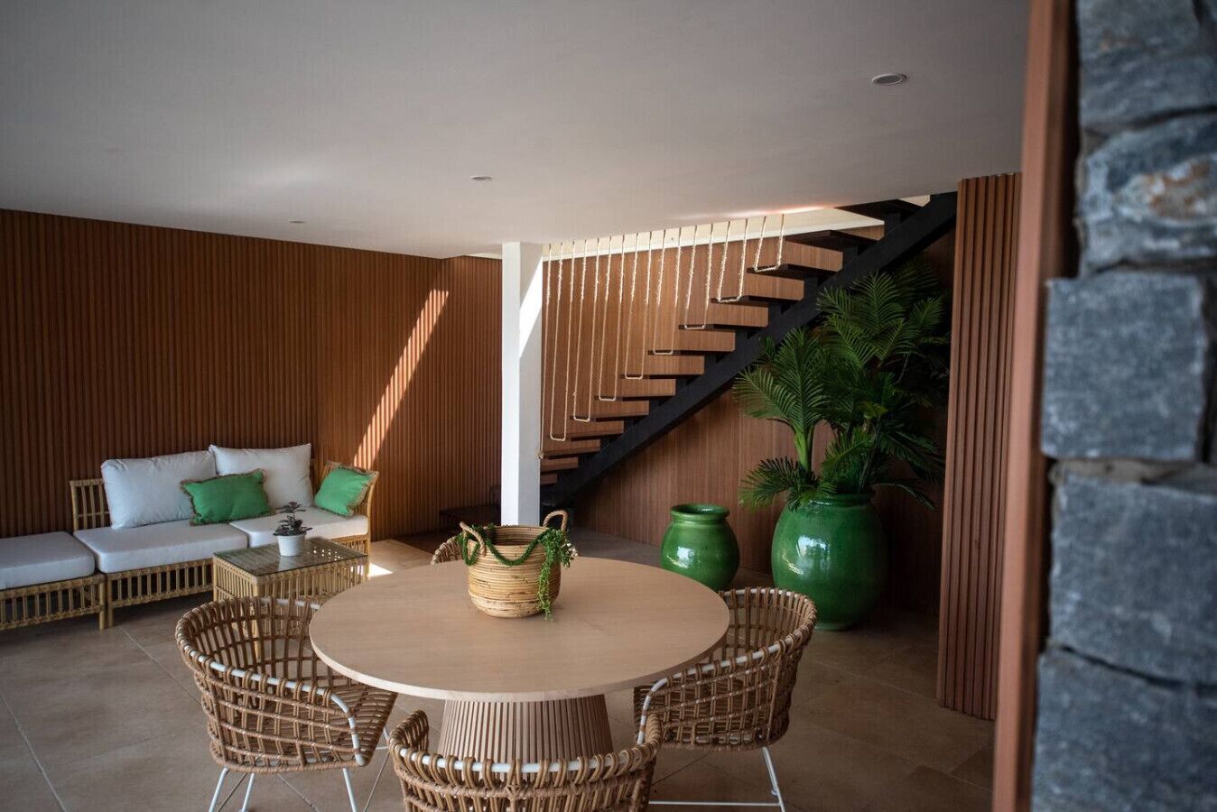 A modern interior with a round wooden table, wicker chairs, a white sofa with green cushions, and a staircase with wooden steps.