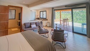 A modern hotel room with a bed, sofa, chairs, a small kitchen area, and a sliding glass door to a balcony.
