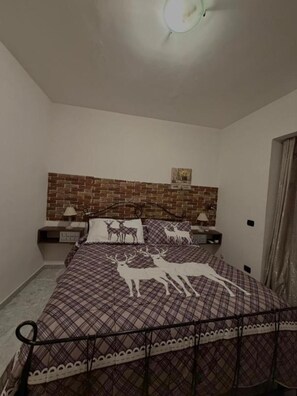Une chambre Ă coucher avec une tĂȘte de lit ornĂ©e dâun motif en briques, un lit recouvert dâune courtepointe Ă carreaux dĂ©corĂ©e de motifs de cerfs, deux tables de chevet avec des lampes et un luminaire fixĂ© au mur.