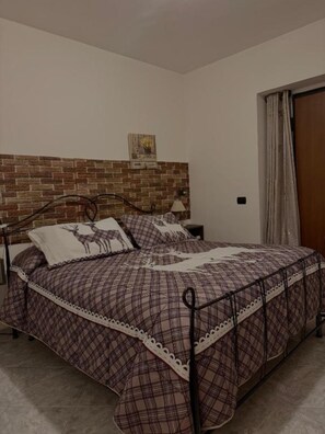 Une chambre Ă coucher avec une tĂȘte de lit ornĂ©e dâun motif en briques, un lit avec une couette Ă carreaux, une table de chevet, une lampe et une porte avec des rideaux.