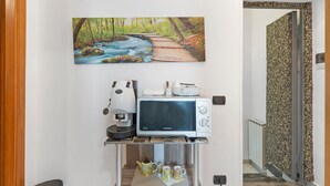 Een keukenhoek met een magnetron, koffieapparaat en theepot op een plank, daarboven een schilderij van een beek in het bos, en een wand met een patroon.