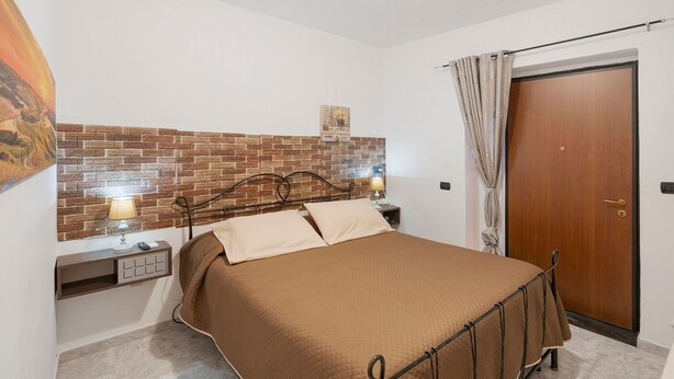 Een slaapkamer met een hoofdeinde van baksteenmotief, een bed met een bruine deken, een nachtkastje met een lamp en een houten deur.