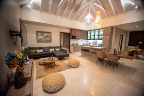 A spacious living area with a sofa, round ottomans, a dining table with chairs, a kitchen in the background, and a ceiling with exposed beams. - Sweet Hill - Villa Rosa (Petite Saline)