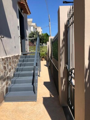 Un étroit sentier mÚne à une entrée fermée par un portail, avec un escalier sur la gauche et un bùtiment sur la droite.
