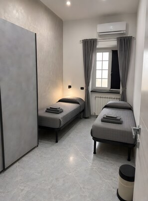 Une chambre de taille modeste, Ă©quipĂ©e de deux lits simples, dâun radiateur, dâun appareil de climatisation, dâune fenĂȘtre avec des rideaux et dâune poubelle.