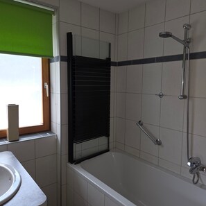 Une salle de bain avec une baignoire, une douche, une fenĂȘtre avec des stores et un porte-serviettes.