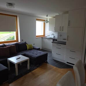 A modern living room with a sofa, a small table, and a kitchen area with cabinets and a window.