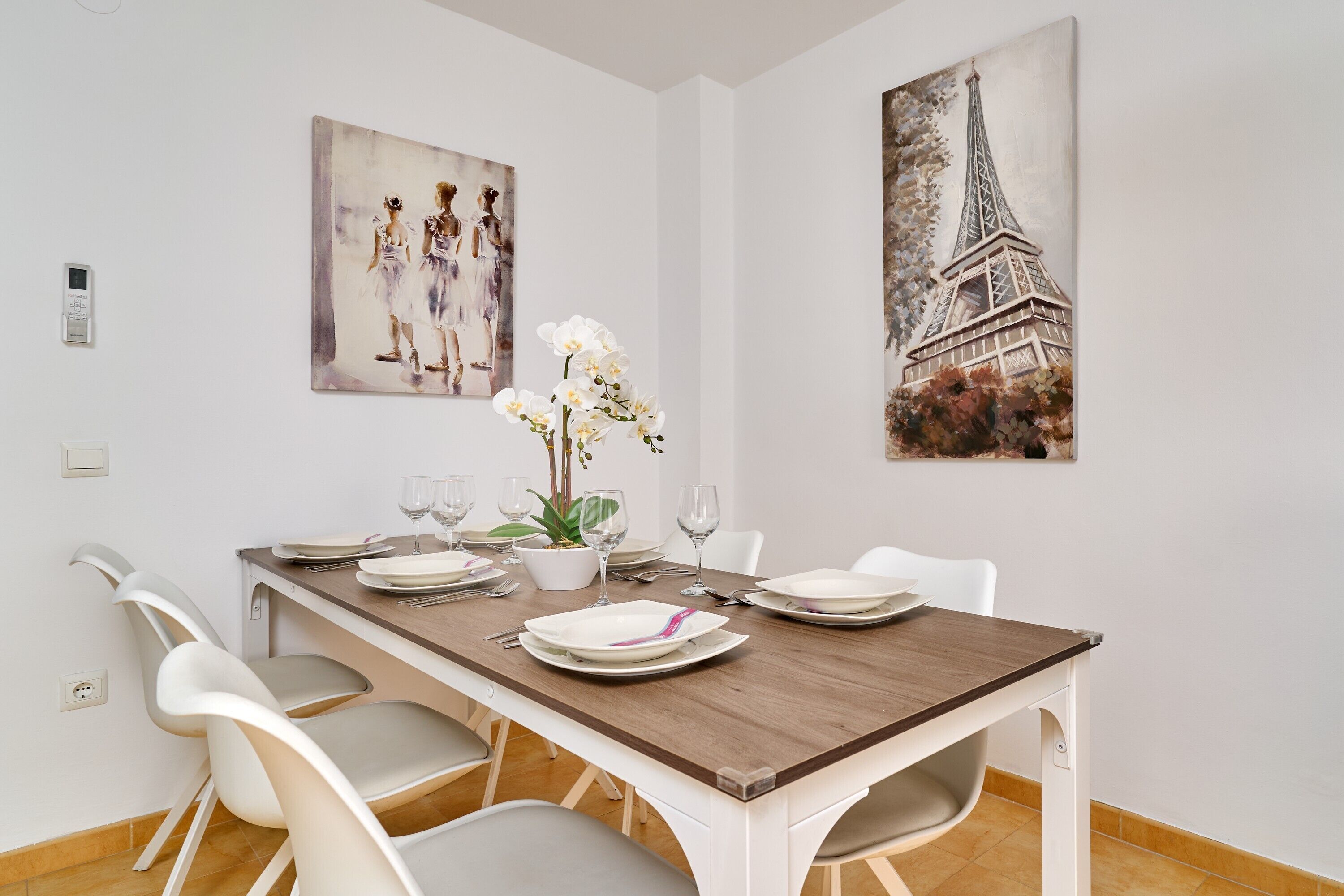 Un coin repas avec une table en bois, des chaises blanches et des œuvres d’art représentant la Tour Eiffel et deux personnages.