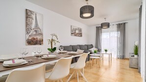 A modern dining area with a table set for four, a grey sofa, and a view of the Eiffel Tower through sheer curtains. - Villa Oasis del Sol II – Pool – Sea Views (Urbanización Cerro Garbancito)