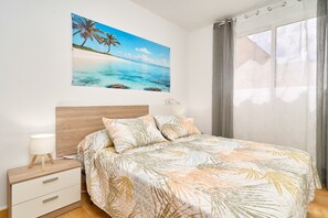 Een slaapkamer met een bed, nachtkastje, lamp, schilderij van een strand en een raam met gordijnen.