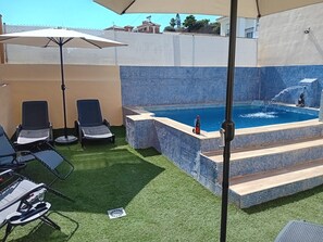 En swimmingpool med flisegrunde trin, to liggestole, et bord med et glas og en parasol.