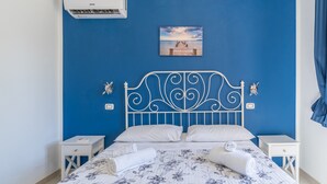 A bedroom with a blue wall, a white metal headboard, two white nightstands, a bed with floral bedding, and a painting of a pier. - Apartment 'Milu' Poolside, Wi-Fi and Air Conditioning (Arenella)