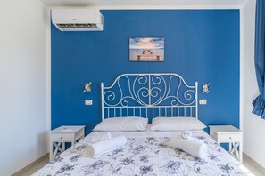 Ein Schlafzimmer mit blauer Wand, einem weißen Metall-Kopfteil, zwei weißen Nachttischen, einem Bett mit floralem Bettzeug und einem Gemälde eines Piers.