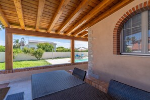 Eine überdachte Terrasse mit Holzboden, Gartenmöbeln und Blick auf einen Swimmingpool sowie benachbarte Häuser.
