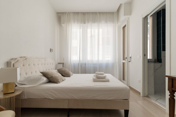 A bedroom with a bed, a bedside table, a window with sheer curtains, and a door leading to a bathroom. - Spacious 100m2 apartment, ideal for up to 6 guests, located on the second floor with lift.Located in the Guastalla district, it is about a 15-minute walk from the M3 Crocetta, M4 Sforza-Policlinico and M1/M4 San Babila metro stops, with numerous surface  (Milan)