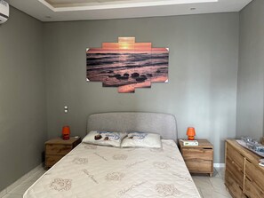 Une chambre à coucher comprenant un lit, deux tables de chevet, une commode en bois et un tableau représentant un coucher de soleil, accroché au mur.