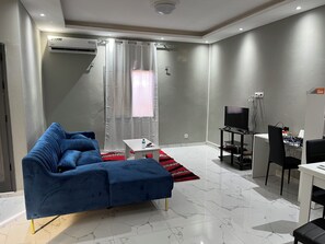 Un salon moderne avec un canapé bleu, une petite table blanche, un téléviseur et un coin repas avec des chaises.