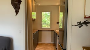 Un estrecho pasillo con suelo de madera, una ventana con vistas a la vegetación, una zona de cocina con armarios y un refrigerador, y un adorno fijado a la pared.