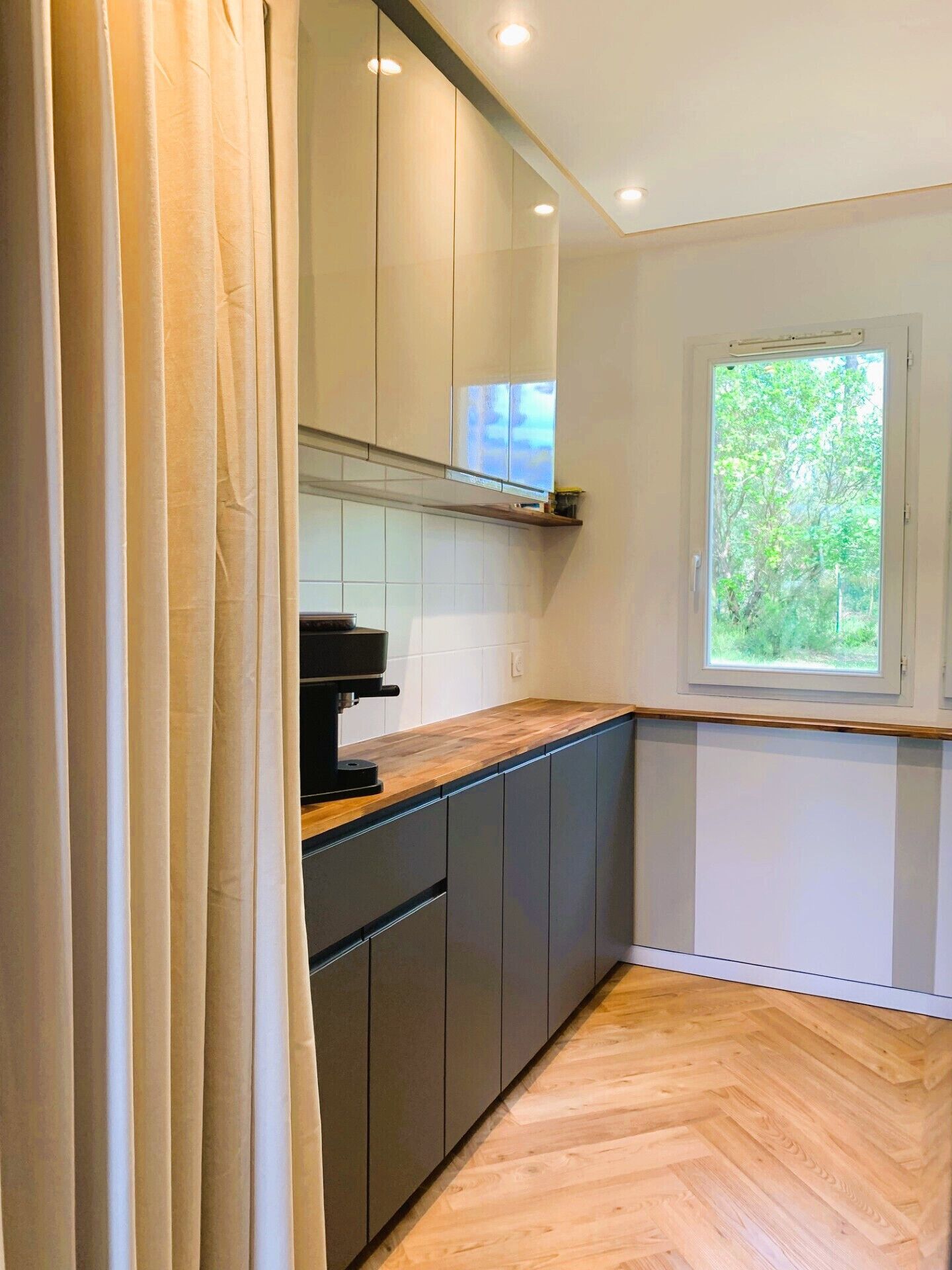 Una cocina moderna con suelo de madera en patrón de espiga, armarios blancos y ventana con vistas a zonas verdes.