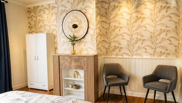 Ein Schlafzimmer mit einem Bett, zwei Stühlen, einem weißen Kleiderschrank, einer Holzregaleinheit und einer Wand mit einem Blattmuster.