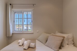 A bedroom with a bed, pillows, a window with curtains, and a view of buildings outside. - #318 Spacious 2BR Apartment | Bright Interiors (Albufeira)