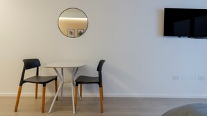 Una habitación con una mesa blanca, dos sillas negras, un espejo redondo y un televisor de pantalla plana montado en la pared.