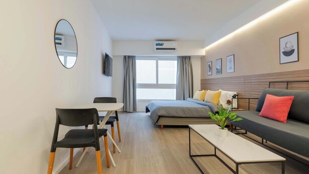 Una habitación de hotel moderna con cama, sofá, mesa de comedor, sillas, un espejo y una planta en maceta.