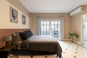 A bedroom with a bed, a nightstand, a lamp, two framed pictures on the wall, a window with curtains, and a potted plant. - Leonardo Da Vinci Aparts 2G by MOT Rentals (Buenos Aires)