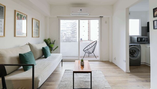 A living room with a sofa, a coffee table, a chair, and a washing machine in the adjacent room. - MOT Rentals + Arevalo 2300 - 3 pax Loft Palermo Hollywood (Buenos Aires)