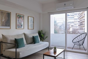 Ein modernes Wohnzimmer mit einer Couch, einem Couchtisch und einem Sessel. An den Wänden hängen gerahmte Kunstwerke und ein großes Fenster bietet Blick auf Gebäude.