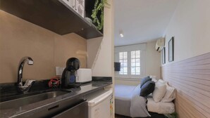 Een compacte keuken met een spoelbak, koffiezetapparaat en een kleine koelkast. In de aangrenzende kamer is een bed met kussens en een plant te zien.