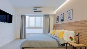 Ein modernes Schlafzimmer mit Bett, montiertem Fernseher, Klimaanlage, einem Fenster mit Vorhängen und gerahmten Bildern an der Wand.